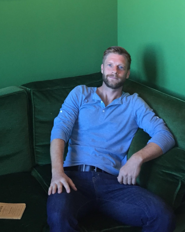 Scott on green couch with blue shirt and blue pants smiling at camera